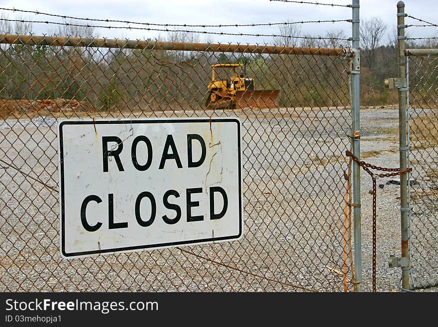 A road closed sign fastened to a chain link fence, a bulldozer in the background