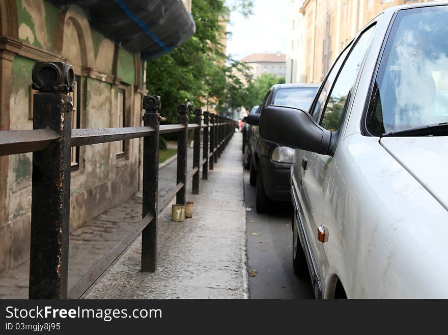 Parked cars in the city. Fence is between the road and the sidewalk. Old houses near to the sidewalk. Parked cars in the city. Fence is between the road and the sidewalk. Old houses near to the sidewalk.