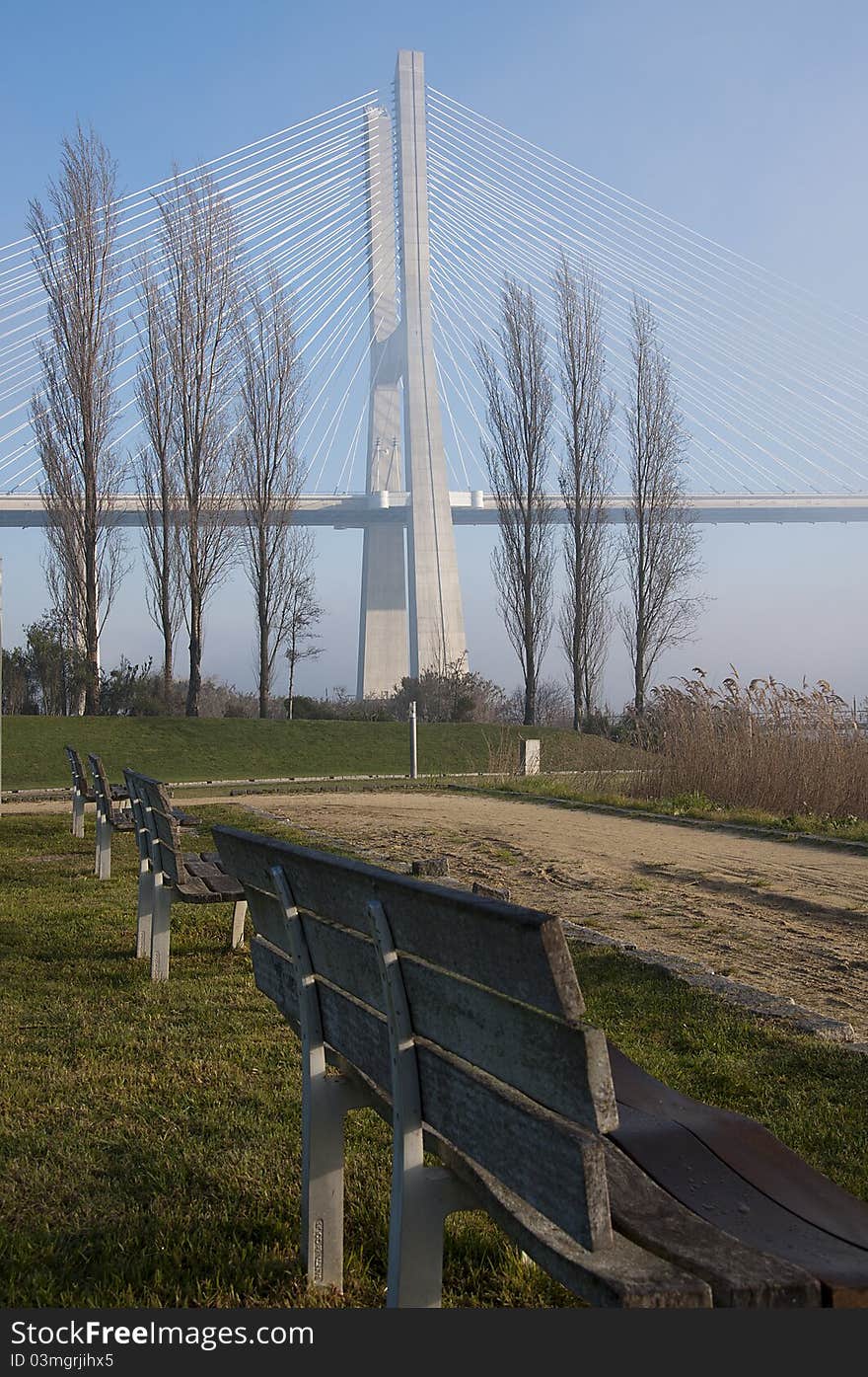 A row of benches lay on a park with a view from several trees and a bridge. A row of benches lay on a park with a view from several trees and a bridge