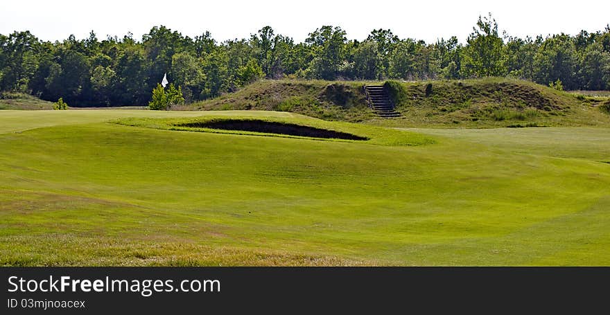 Golf green showing flag pole and green side bunker. In the background is an the next hole’s tee box. The golf course is an old world links style course. Golf green showing flag pole and green side bunker. In the background is an the next hole’s tee box. The golf course is an old world links style course.