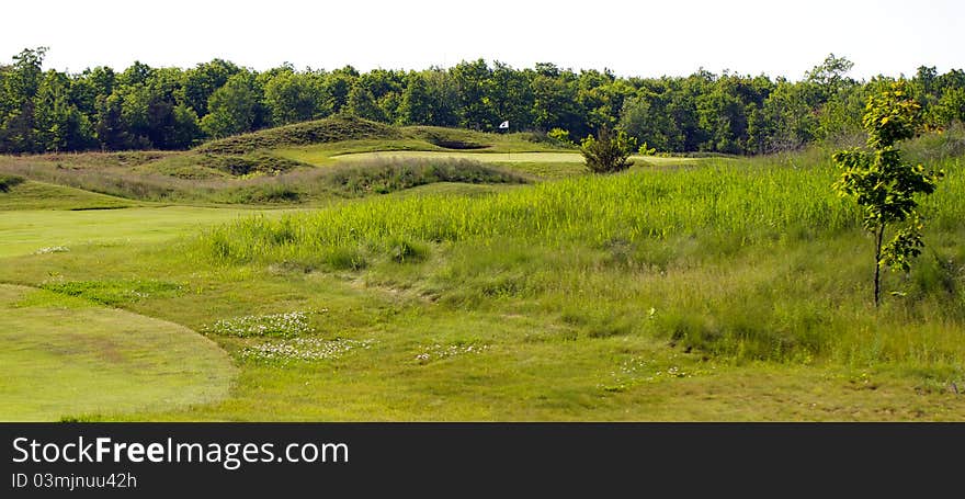 Golf green showing flag pole and green side bunker. In the background is an the next hole’s tee box. The golf course is an old world links style course. Golf green showing flag pole and green side bunker. In the background is an the next hole’s tee box. The golf course is an old world links style course.