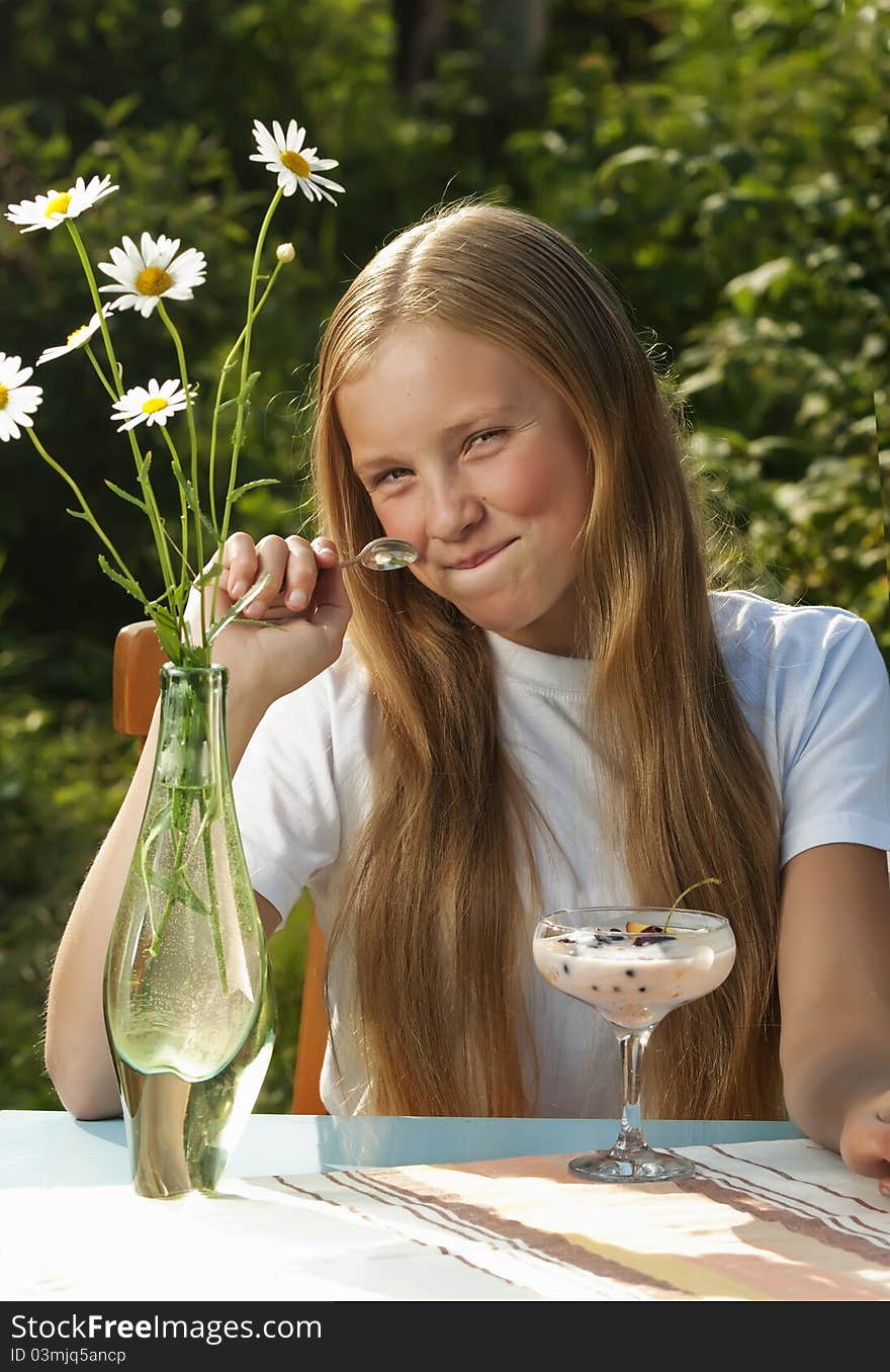The girl eats berry yogurt in a garden in the summer. On a table there is a vase with camomiles. The girl eats berry yogurt in a garden in the summer. On a table there is a vase with camomiles