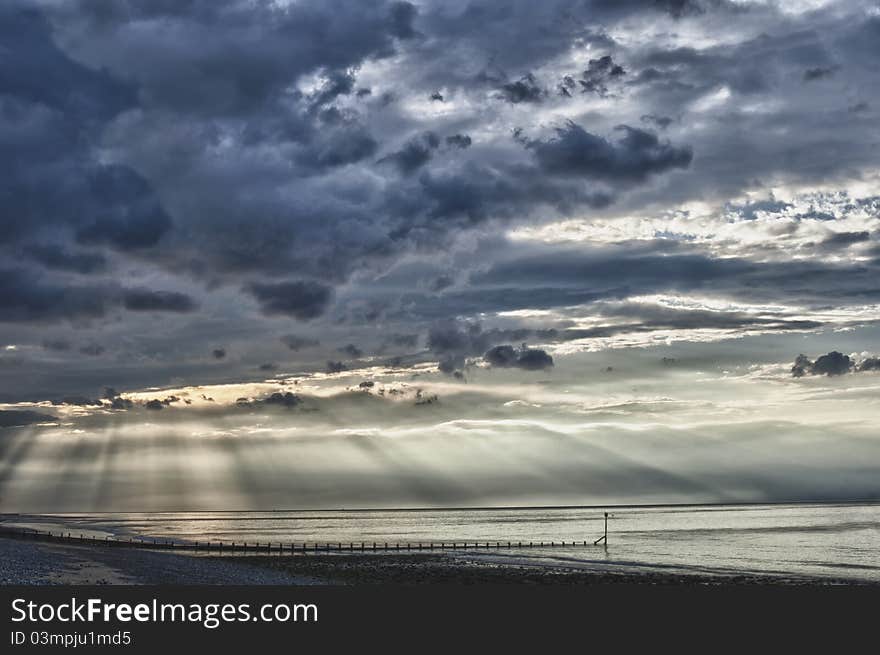 Suns rays bursting through clouds on sea, at near sunset. Breakwater and beach in foreground. HDR image. Suns rays bursting through clouds on sea, at near sunset. Breakwater and beach in foreground. HDR image