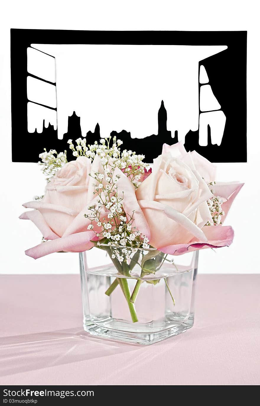 Beautiful pink roses in a square vase on a table at an open window. Window is a cut-out silhouette of a large London-like city. Table is covered with a pink cloth. Beautiful pink roses in a square vase on a table at an open window. Window is a cut-out silhouette of a large London-like city. Table is covered with a pink cloth.