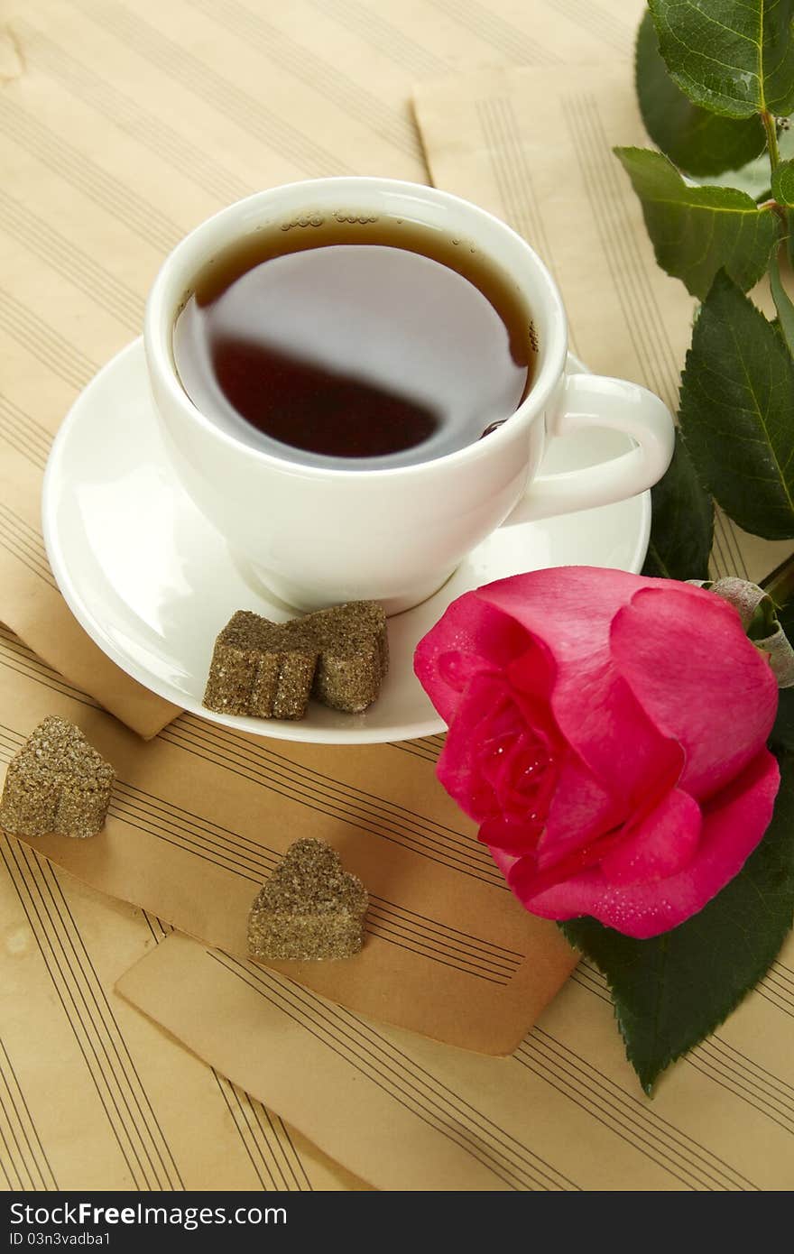 White cup of tea next to sugar and is a beautiful red rose lying on sheets of old paper sheet music. White cup of tea next to sugar and is a beautiful red rose lying on sheets of old paper sheet music