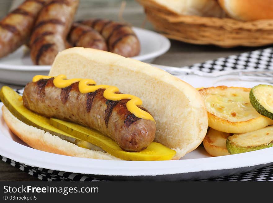 Grilled bratwurst with mustard and pickles on a bun and fried squash. Grilled bratwurst with mustard and pickles on a bun and fried squash