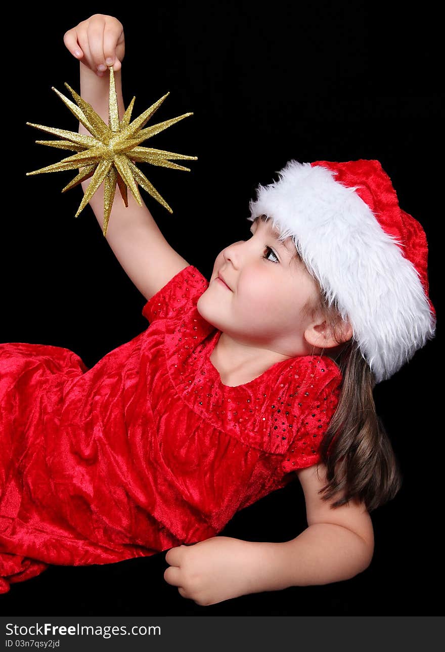 Little girl in Santa hat laying down and looking up at the golden Chritsmas ornament in her hand. isolated on black. Little girl in Santa hat laying down and looking up at the golden Chritsmas ornament in her hand. isolated on black