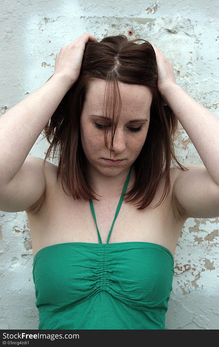 A young woman in a green top stands, playing with her hair, in front of a white-painted wall. A young woman in a green top stands, playing with her hair, in front of a white-painted wall.