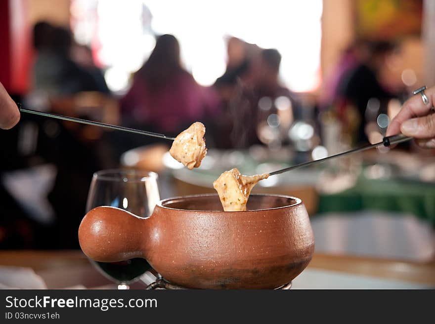 Two people serving fondeau from a ceramic hot bowl in a restaurant and a cup of wine behind, with other people having a meal in the blury background.