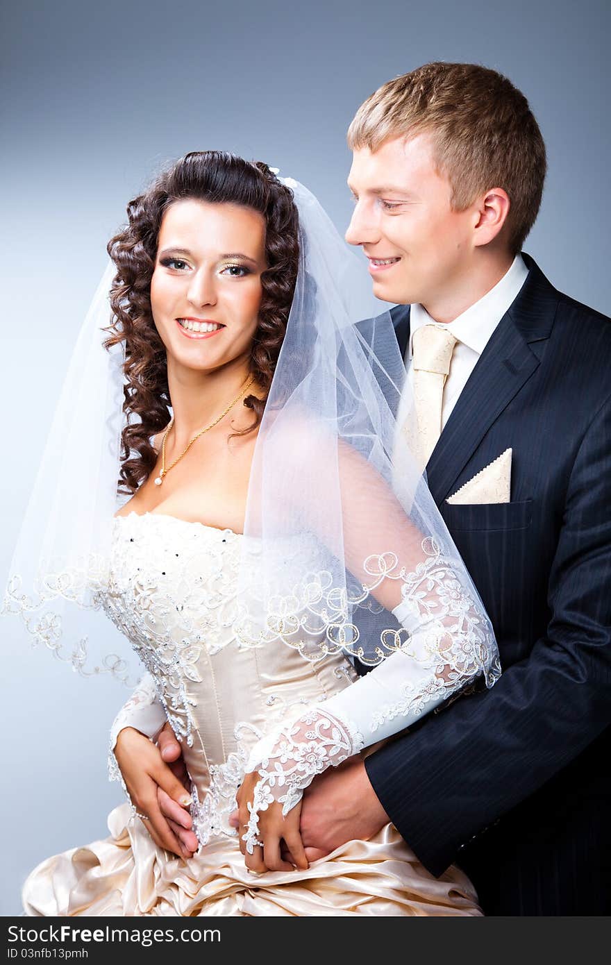 Studio portrait of young elegant enamoured just married groom looking at beautiful bride and embracing on blue background. Studio portrait of young elegant enamoured just married groom looking at beautiful bride and embracing on blue background