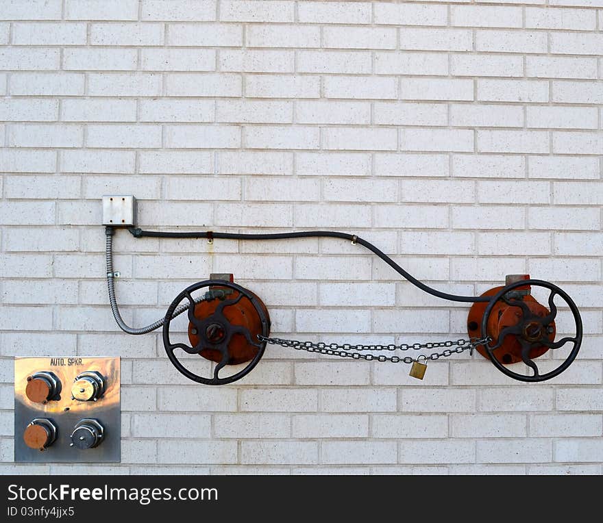 A unique vehicle-like design created from conduits, two round fire hose water shut off valves and a safety chain against the side of a white brick building. A unique vehicle-like design created from conduits, two round fire hose water shut off valves and a safety chain against the side of a white brick building.