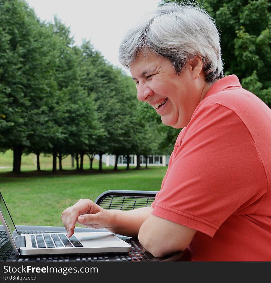 Middle-aged woman browsing on laptop at table outside with trees. Middle-aged woman browsing on laptop at table outside with trees