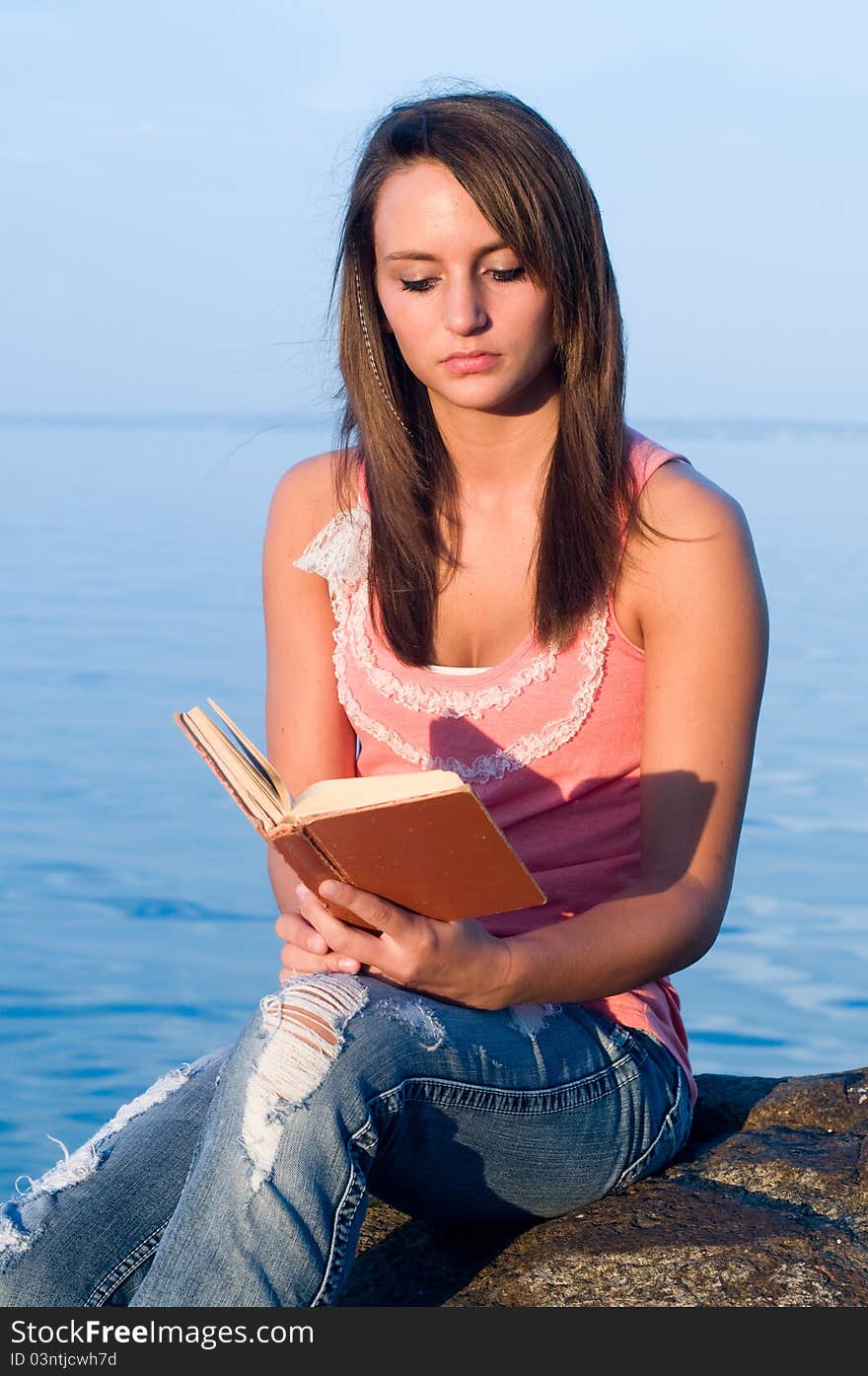 Woman reading on a rock at the edge of an ocean or seaside or body of water.