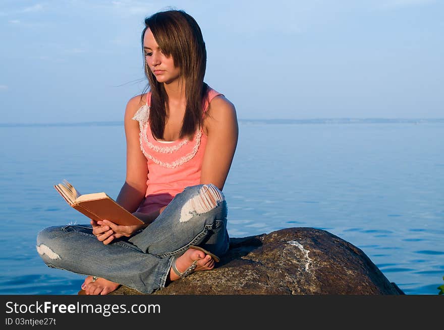 Woman reading on a rock at the edge of an ocean or seaside or body of water. She is at peace and enjoying her downtime.