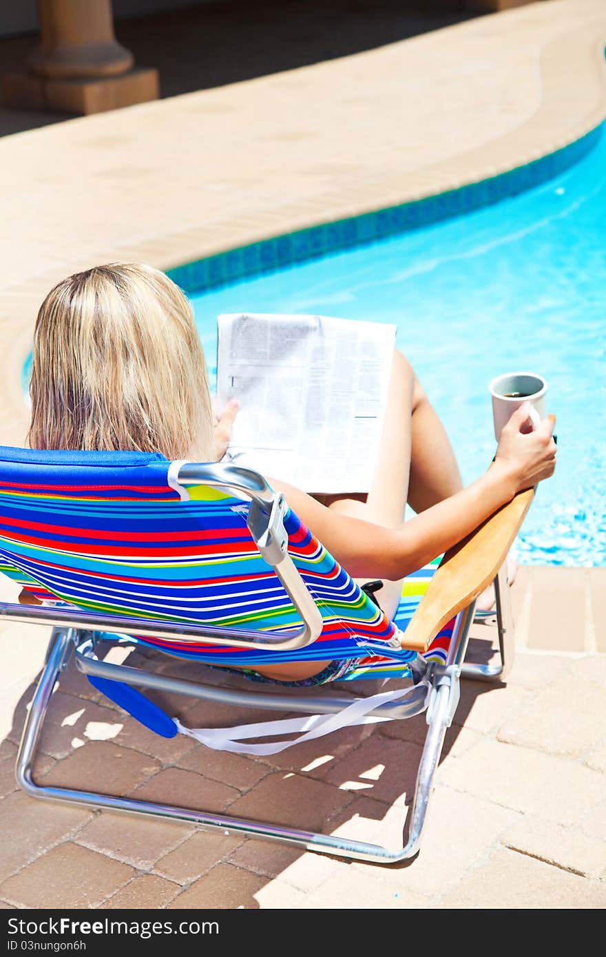 The woman is holding cup of coffee and reading newspaper by the pool. The woman is holding cup of coffee and reading newspaper by the pool