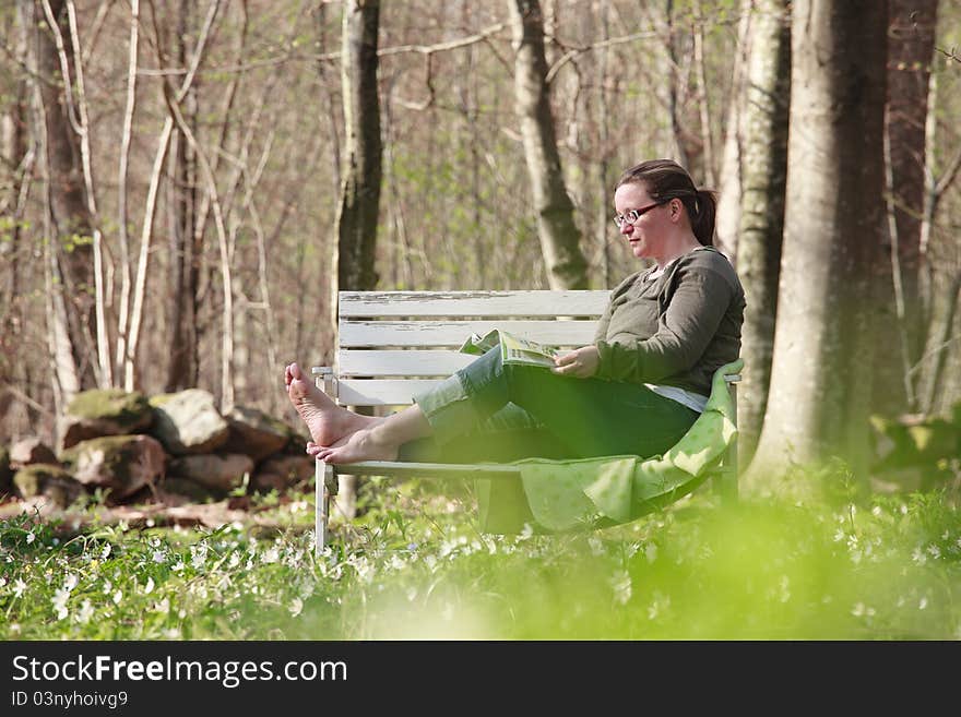 Woman sitting on a sofa in the wood in springtime. She is reading a magazine in the sunlight. Woman sitting on a sofa in the wood in springtime. She is reading a magazine in the sunlight