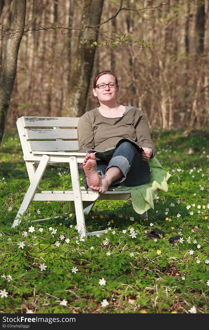 Woman sitting on a sofa in the wood in springtime. She is reading a magazine in the sunlight. Woman sitting on a sofa in the wood in springtime. She is reading a magazine in the sunlight