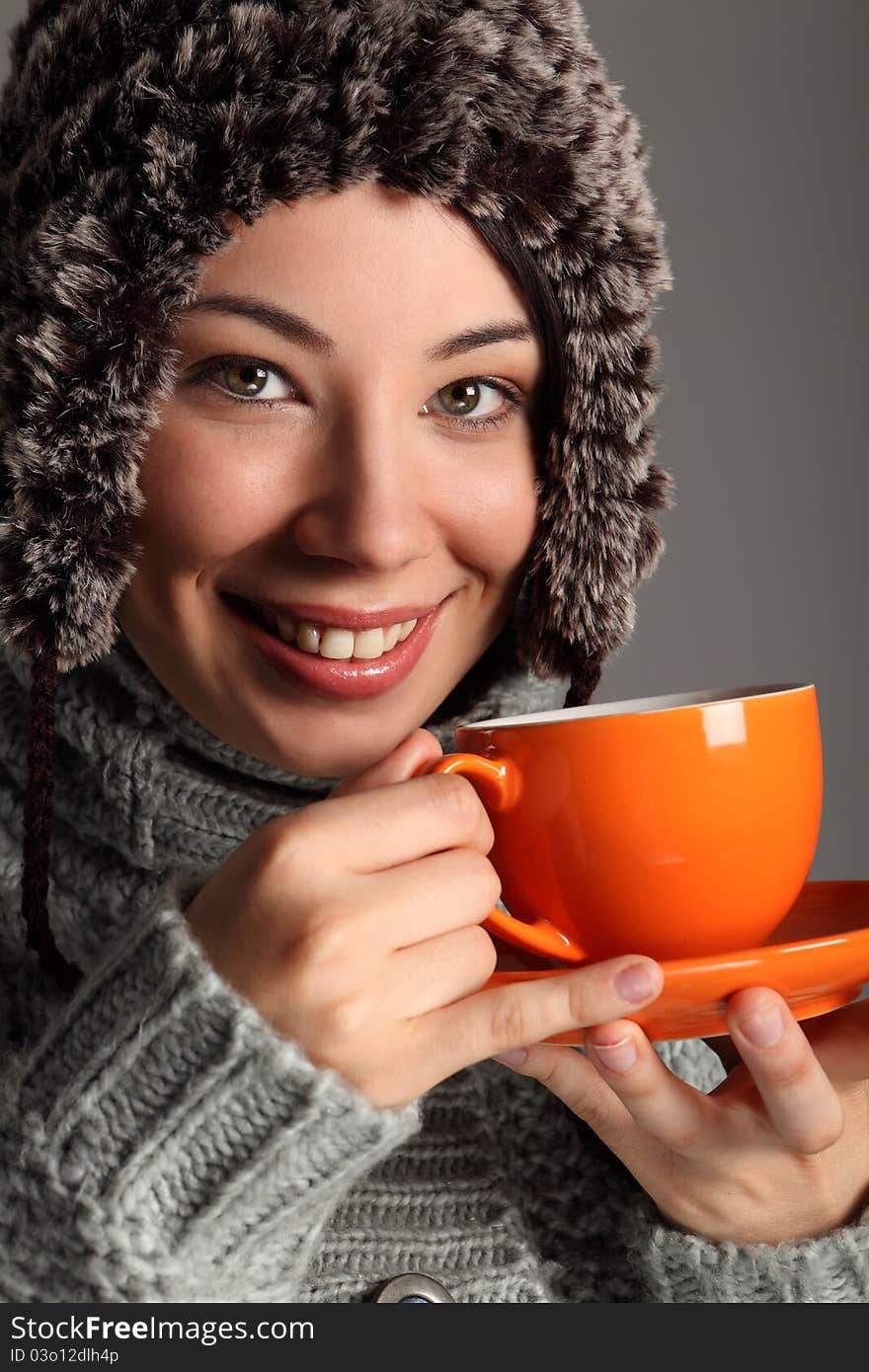 Beautiful smile from young woman wearing thick wool sweater and furry winter hat, drinking tea from orange cup. Shot against a grey background. Beautiful smile from young woman wearing thick wool sweater and furry winter hat, drinking tea from orange cup. Shot against a grey background.