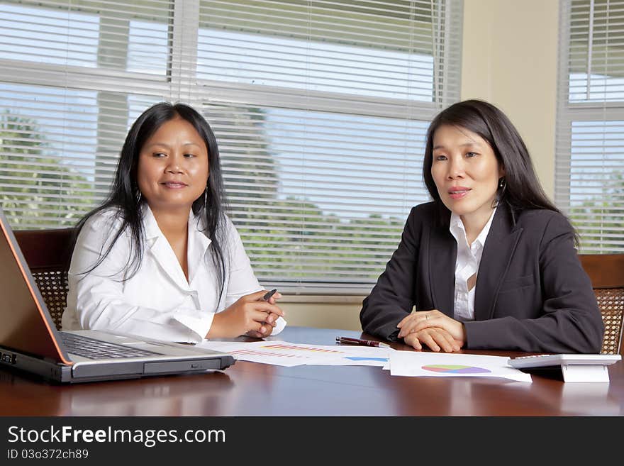 Two Asian Women at desk with laptop charts, graphs on desk.