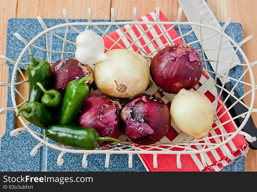 Jalapeno peppers, red onions, yellow onions, and a head of garlic rest in a wire basket on top of a cutting board next to a chef knife and red stripe towel. Jalapeno peppers, red onions, yellow onions, and a head of garlic rest in a wire basket on top of a cutting board next to a chef knife and red stripe towel.