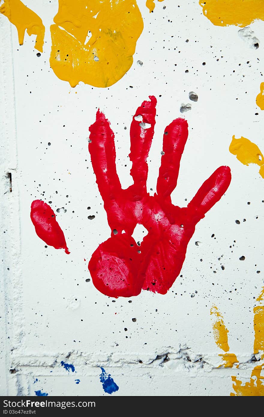 A child's handprint in red paint on a white stone wall. Other paint spots in yellow and blue are also seen.