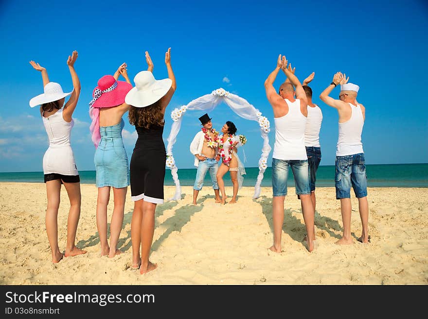 Groom with bride wearing lei, standing under archway on beach and their friends applauding for joy. Groom with bride wearing lei, standing under archway on beach and their friends applauding for joy