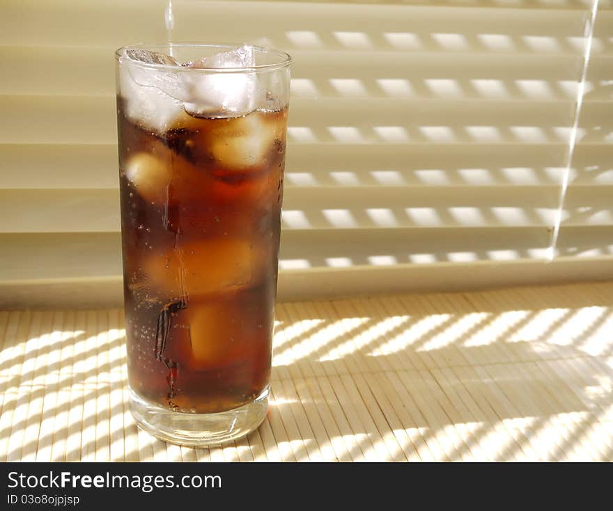 An ice cold fizzy drink with ice in a tall glass on a cream blind background on a sunny day. An ice cold fizzy drink with ice in a tall glass on a cream blind background on a sunny day