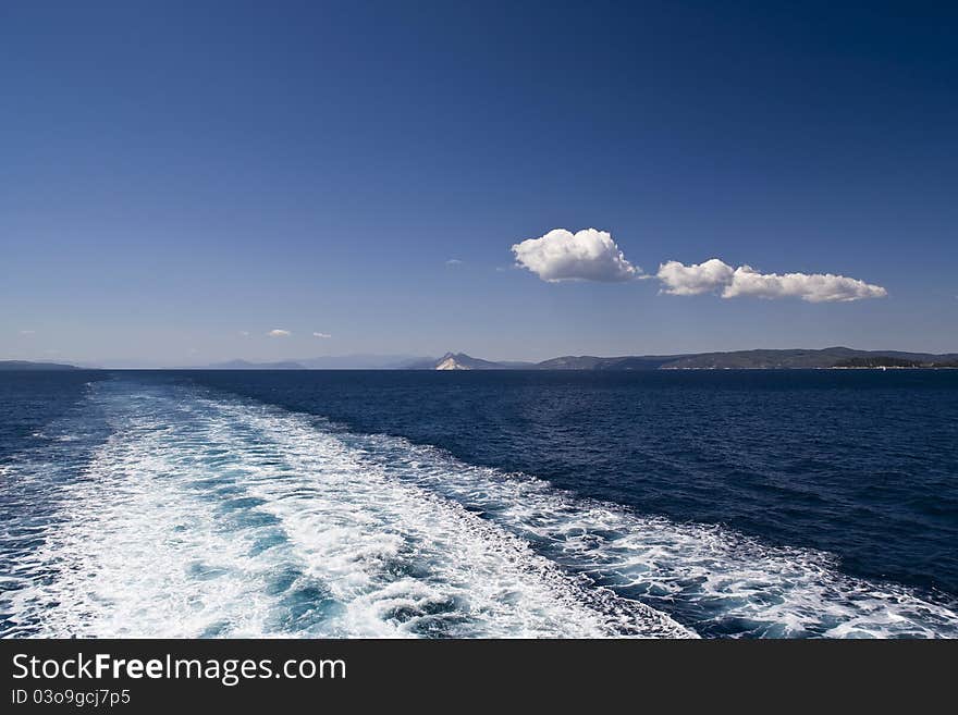 View from a ferryboat on the way to Greek island Alonissos in the Mediterranean Sea.Beautiful seascape with dominant blue colour of sea and sky contrasted with a couple of full well defined white clouds and white water trail. View from a ferryboat on the way to Greek island Alonissos in the Mediterranean Sea.Beautiful seascape with dominant blue colour of sea and sky contrasted with a couple of full well defined white clouds and white water trail.