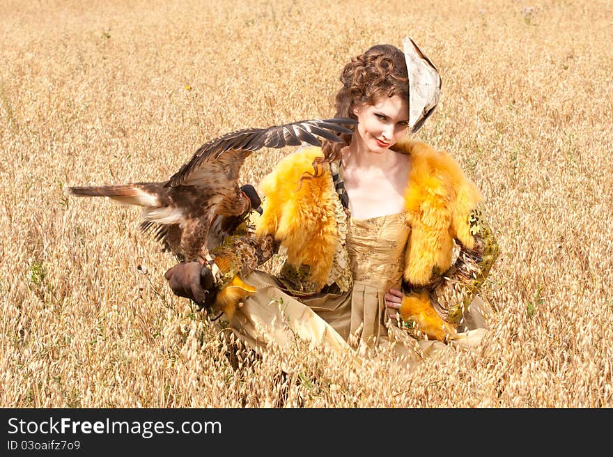 The woman in a beautiful old style dress with falcon has a rest before hunting in yellow field. The woman in a beautiful old style dress with falcon has a rest before hunting in yellow field.