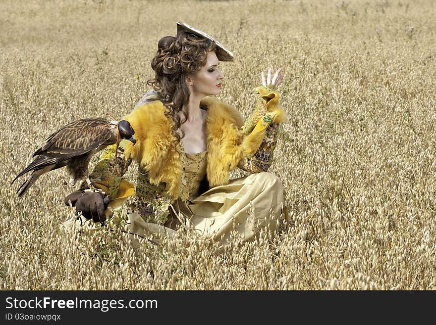 The woman in a beautiful old style dress with falcon has a rest before hunting in yellow field. The woman in a beautiful old style dress with falcon has a rest before hunting in yellow field.