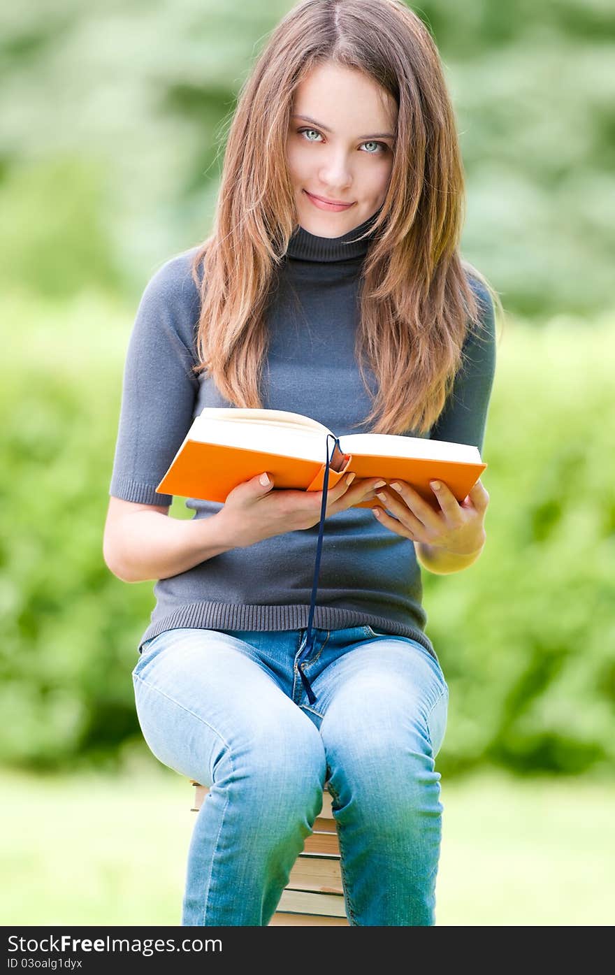 Beautiful and happy young student girl sitting on pile of books, holding book in her hands, smiling and looking into the camera. Summer or spring green park in background. Beautiful and happy young student girl sitting on pile of books, holding book in her hands, smiling and looking into the camera. Summer or spring green park in background