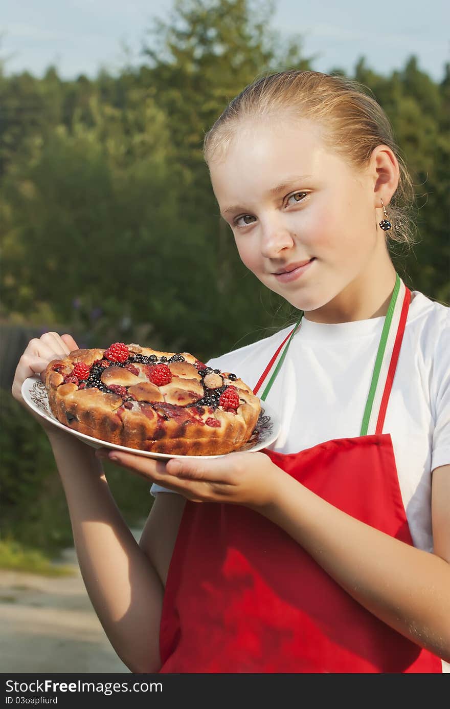 The girl the teenager in a red apron holds in a hand a pie from fresh berries against summer wood. The girl the teenager in a red apron holds in a hand a pie from fresh berries against summer wood