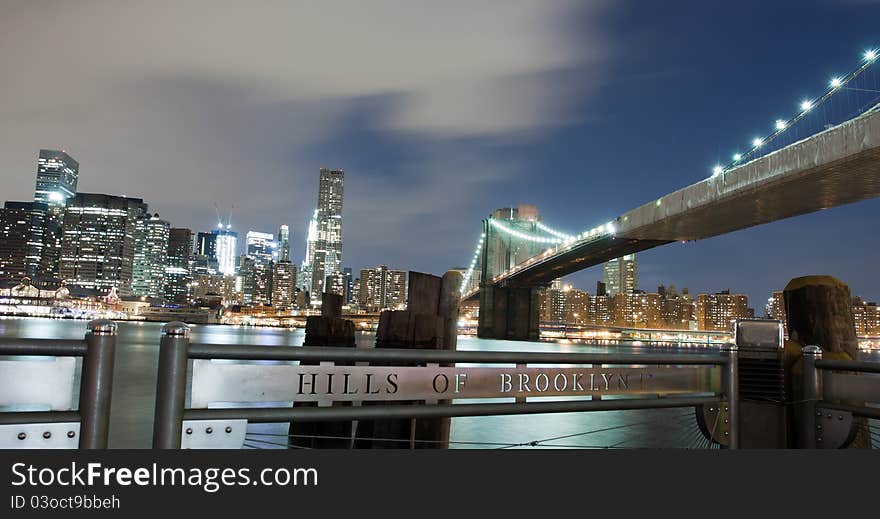 New York City Brooklyn Bridge skyline with skyscrapers over Hudson River illuminated with lights at dusk.