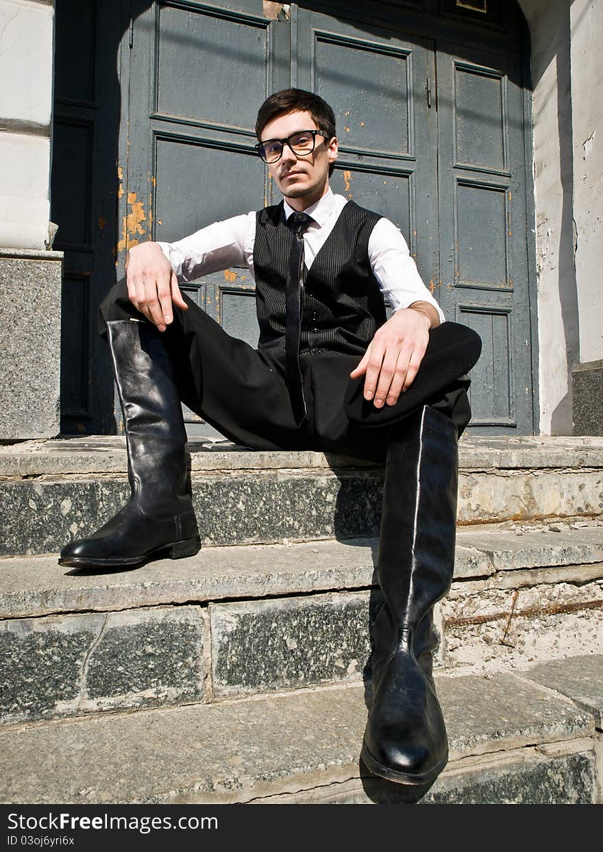 Young man in eyeglass and boots in vintage style is sitting against the background old door. Young man in eyeglass and boots in vintage style is sitting against the background old door