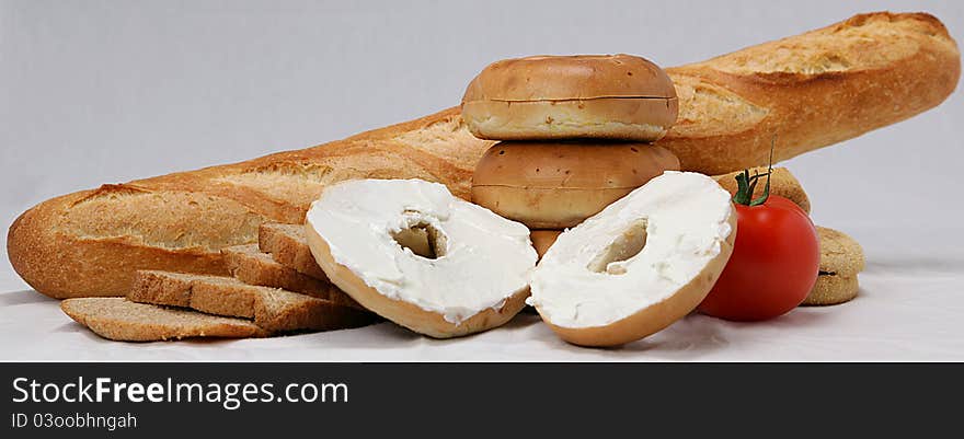 Bagel opened and spread with cream cheese in front of a variety of bread and a tomato. Bagel opened and spread with cream cheese in front of a variety of bread and a tomato.