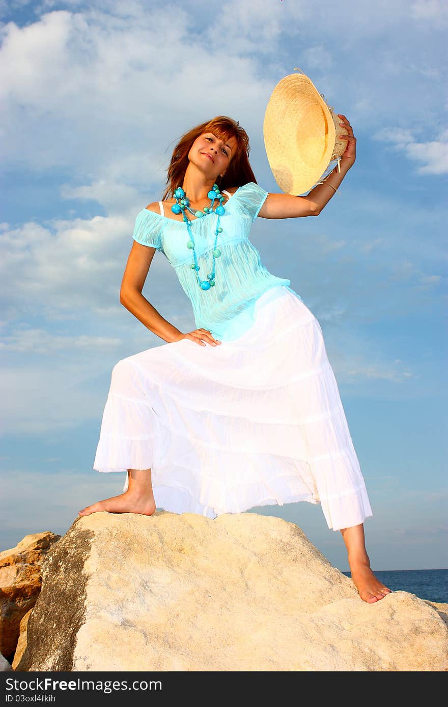 Attractive woman holding his hat (squash) in a hand step on a rock with cloudy sky and blue sea (ocean) background. Attractive woman holding his hat (squash) in a hand step on a rock with cloudy sky and blue sea (ocean) background