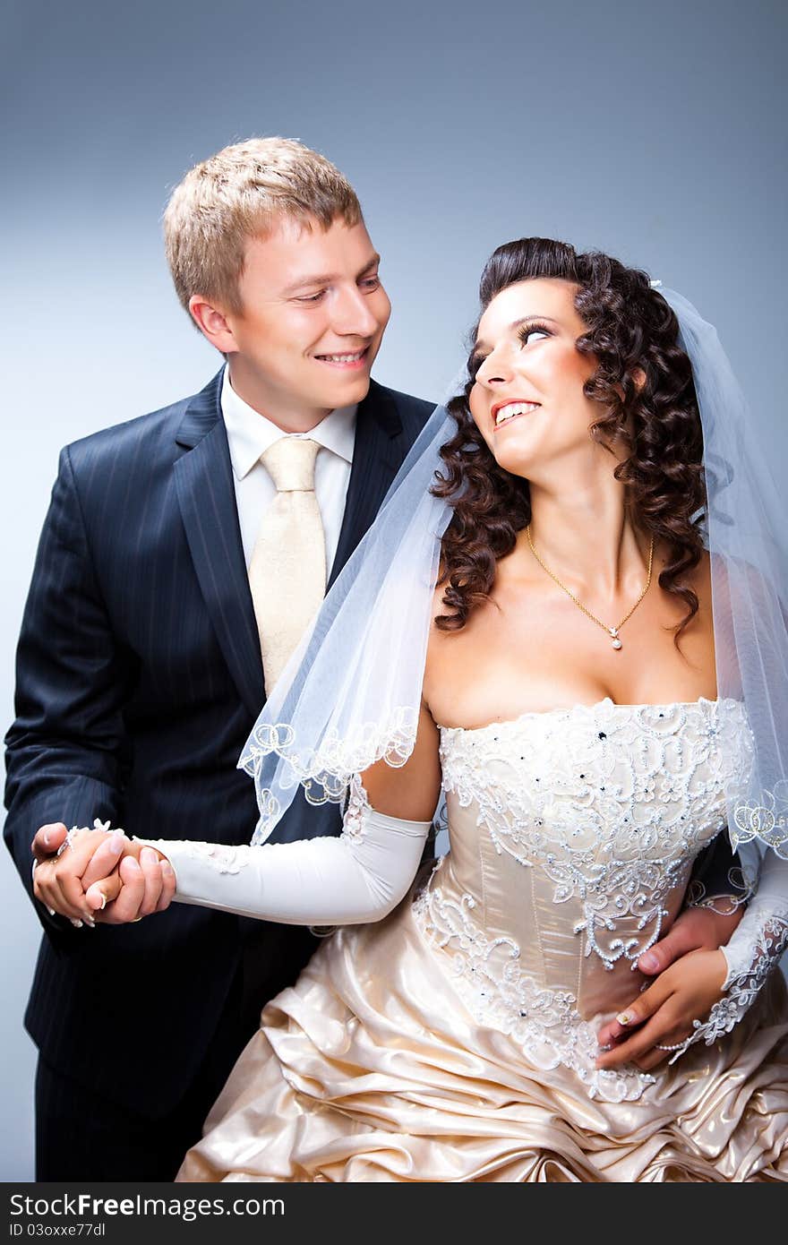 Studio portrait of young elegant enamored just married bride and handsom groom looking to each other and embracing on gray background. Studio portrait of young elegant enamored just married bride and handsom groom looking to each other and embracing on gray background