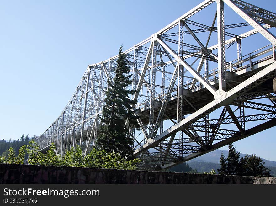 A closeup of an image of the historic Bridge of the Gods in Cascade Locks, Oregon over the Columbia River Gorge with trees in the foreground & blue sky. Selective focus. A closeup of an image of the historic Bridge of the Gods in Cascade Locks, Oregon over the Columbia River Gorge with trees in the foreground & blue sky. Selective focus.