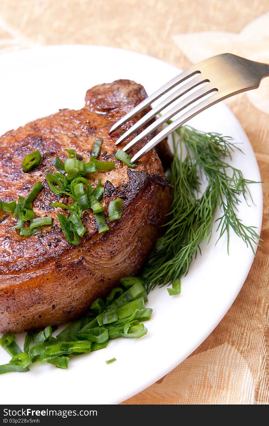 Big juicy well-done piece of meat lying on a white plate decorated with greenery with a fork. Big juicy well-done piece of meat lying on a white plate decorated with greenery with a fork