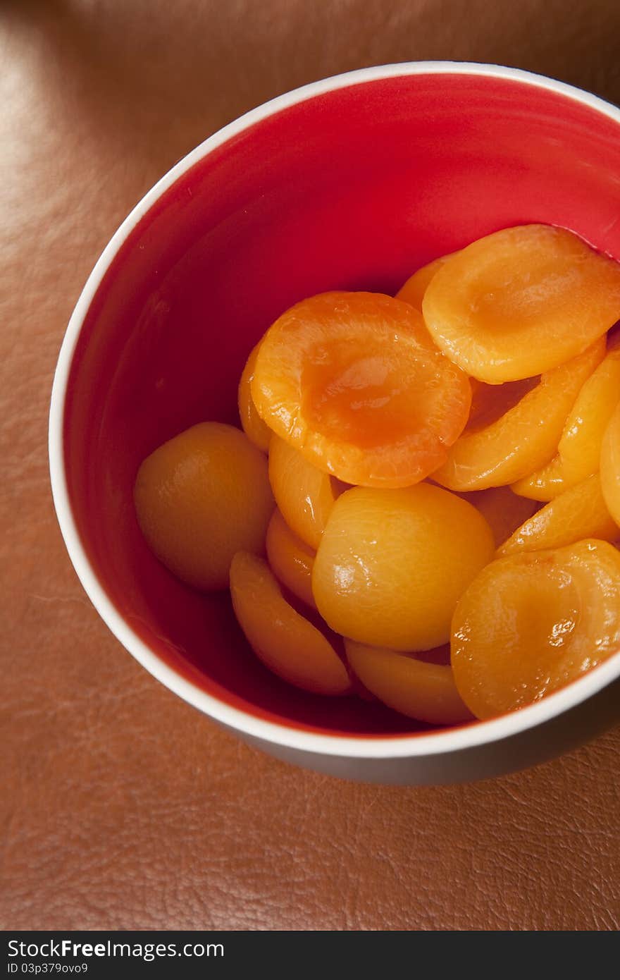 Delicious peaches in syrup sauce in a red bowl, on leather background.