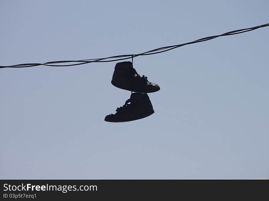 A pair of discarded black shoes hanging from a telephone wire with a blue sky in the background. A pair of discarded black shoes hanging from a telephone wire with a blue sky in the background.