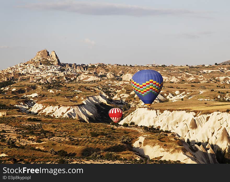 Uchisar backdrop red and white plus blue multi colored hot air balloons navigate a gorge during low level flight, blue sky, wilderness, desert plants, limestone cleft, volcanic region, landscape, crop space and copy area. Uchisar backdrop red and white plus blue multi colored hot air balloons navigate a gorge during low level flight, blue sky, wilderness, desert plants, limestone cleft, volcanic region, landscape, crop space and copy area