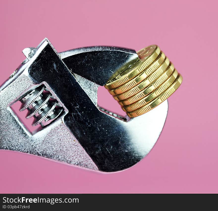 A stack of Gold Dollar Coins being squeezed in a silver adjustable spanner, against a light pastel pink background and asking the questions how tight is you household budget?. A stack of Gold Dollar Coins being squeezed in a silver adjustable spanner, against a light pastel pink background and asking the questions how tight is you household budget?