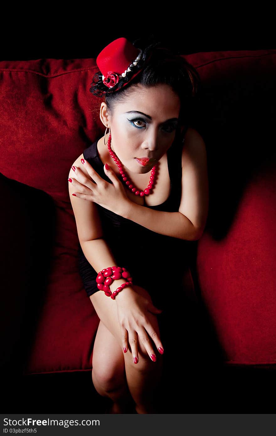 Asian female wearing red hat and red beads looking up towards the camera. Sitting on a red couch on black background.