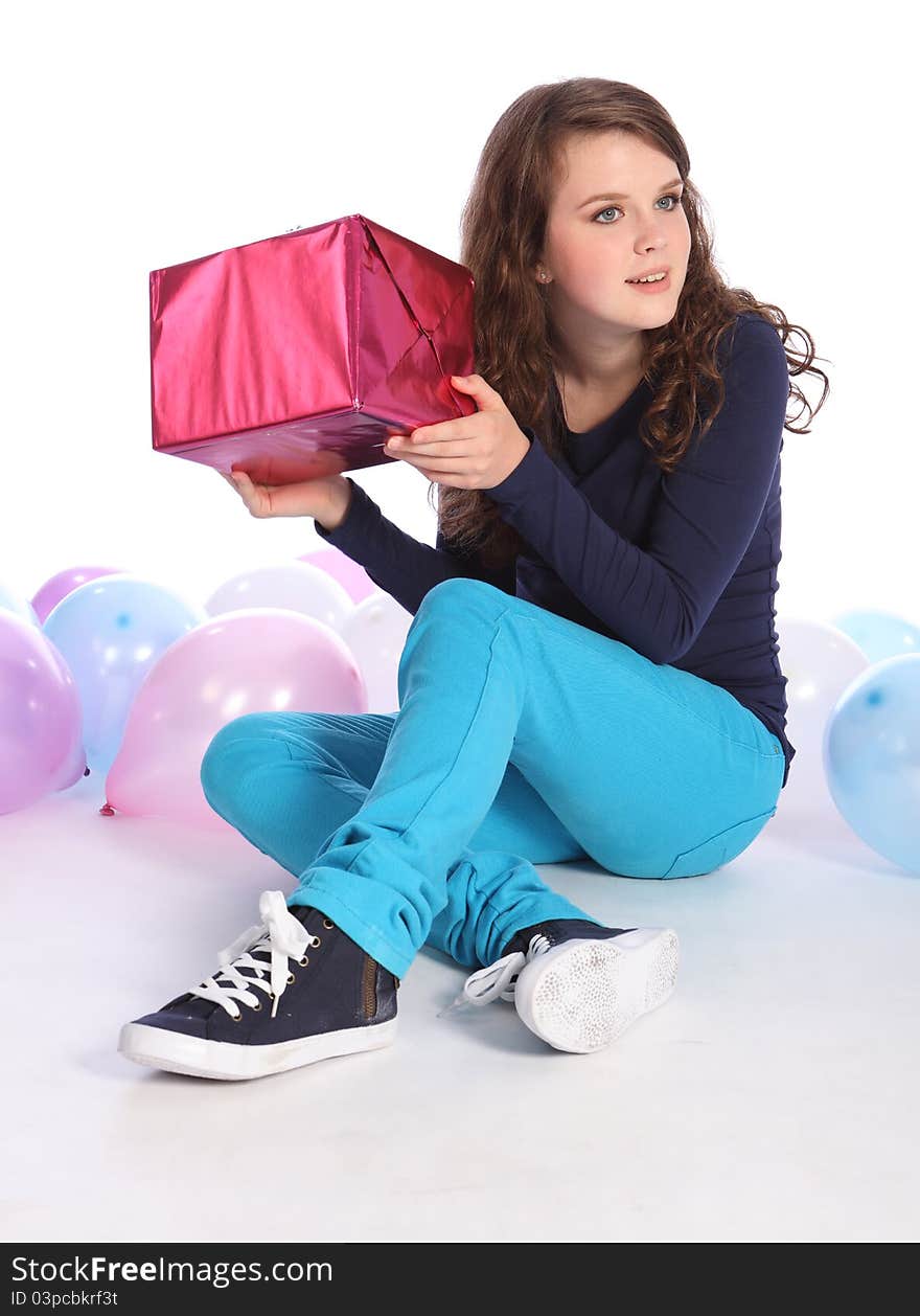 Beautiful teenager girl with bright blue eyes celebrates happy occasion with a surprise birthday present wrapped in pink gift paper, sitting among party balloons. Beautiful teenager girl with bright blue eyes celebrates happy occasion with a surprise birthday present wrapped in pink gift paper, sitting among party balloons.