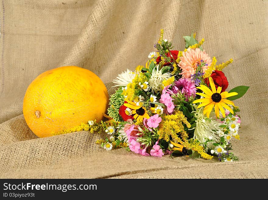 The autumn topic. In the foreground is bouquet of wild and cultivated flowers and orange melon. They lie on the table covered by rough stuff. This staff creates background so. The autumn topic. In the foreground is bouquet of wild and cultivated flowers and orange melon. They lie on the table covered by rough stuff. This staff creates background so.