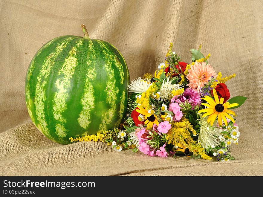 The autumn topic. In the foreground is bouquet of wild and cultivated flowers and green melon. They lie on the table covered by rough stuff. This staff creates background so. The autumn topic. In the foreground is bouquet of wild and cultivated flowers and green melon. They lie on the table covered by rough stuff. This staff creates background so.