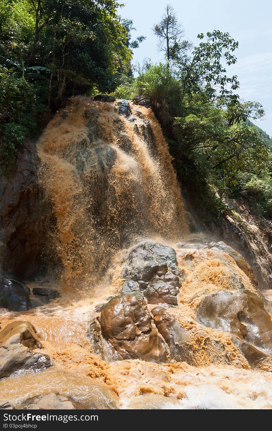 A clayey waterfall with brown water running fast from the hill. One of the stones resembles a human face.