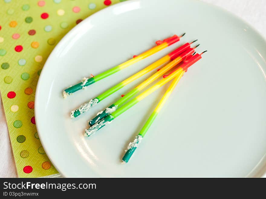 Rainbow birthday candles pulled from the cake and laid on a blue plate. Bits of frosting cling to the drippy, burned, candles. A green polka dot napkin rests below. Rainbow birthday candles pulled from the cake and laid on a blue plate. Bits of frosting cling to the drippy, burned, candles. A green polka dot napkin rests below.