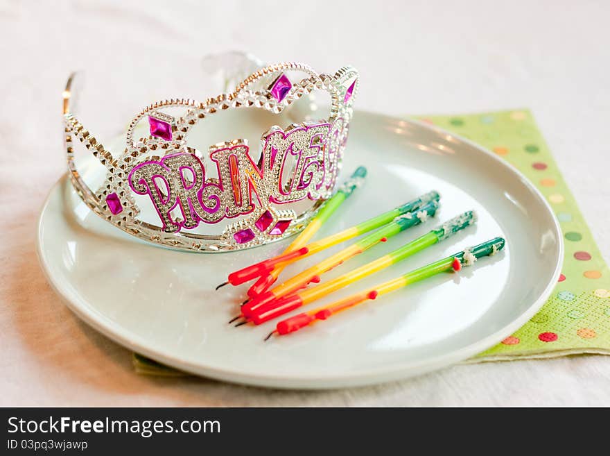 A sparkly rhinestone tiara with silver and purple beads that spells out Princess sits on a blue plate next to used rainbow birthday candles that have been blown out. A sparkly rhinestone tiara with silver and purple beads that spells out Princess sits on a blue plate next to used rainbow birthday candles that have been blown out.